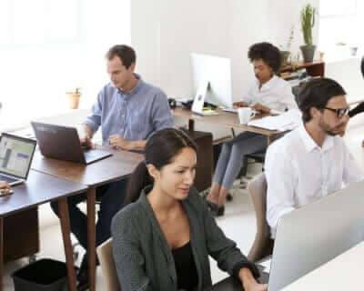 Zwei männliche und zwei weibliche Mitarbeiter sitzen an ihren Schreibtischen in einem hellen Büro und arbeiten an ihren Mac-Desktop-Computern